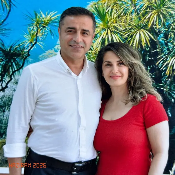 Görselde, Edirne Cezaevinde çekilmiş fotoğraf. Sol tarafta beyaz gömlekli Selahattin Demirtaş, sağ tarafta kırmızı tişörtlü Başak Demirtaş, kameraya bakıp gülümsüyorlar. Arka planda, cezaevi görüş odasındaki tabloda ağaç dalları var. Sol alt köşede dijital yazıyla Haziran 2026 yazılı.