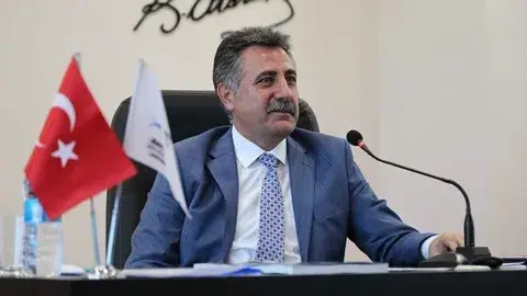 Serdar Sandal bir toplantı salonunda mikrofon önünde otururken görülüyor.